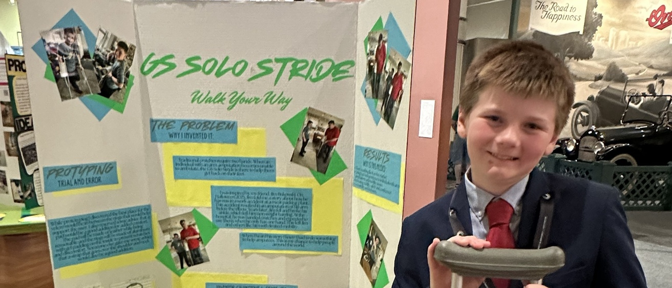 An Iowa student named Gavin standing with his invention and display board at nationals